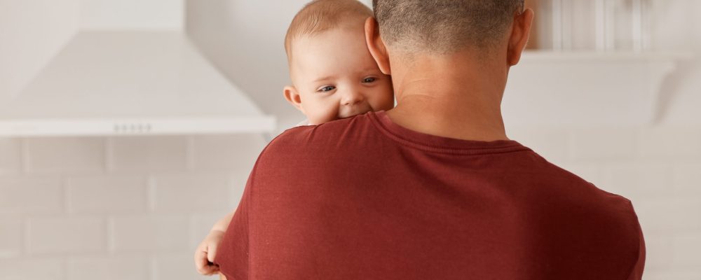 Back view portrait of young adult Caucasian father holding son or daughter, cute baby looking at camera over mans shoulder, posing indoor with kitchen set on background.