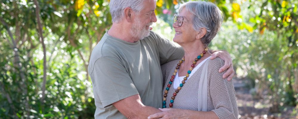 Beautiful white-haired senior couple walking in the woods hug looking in the eyes. Smiling elderly grandparents enjoy healthy lifestyle in public park