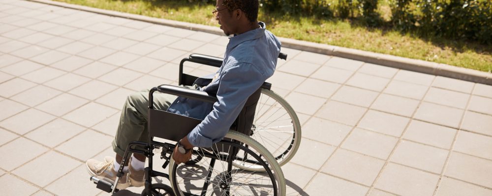 Young Black man with aching legs moving forward in wheelchair over street, rehab concept