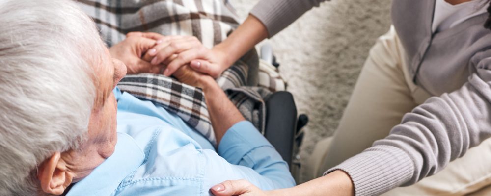 Young supportive female caregiver sitting by senior man in wheelchair and keeping her hand on his shoulder while comforting him
